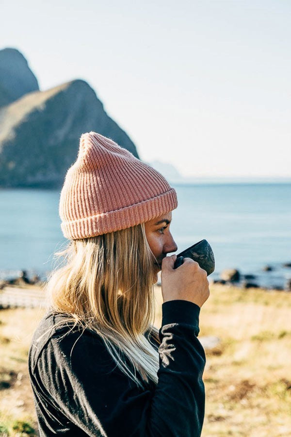 Femme au bonnet rose buvant dans une Kupilka 21 Classic Cup noire sur la côte près des montagnes
