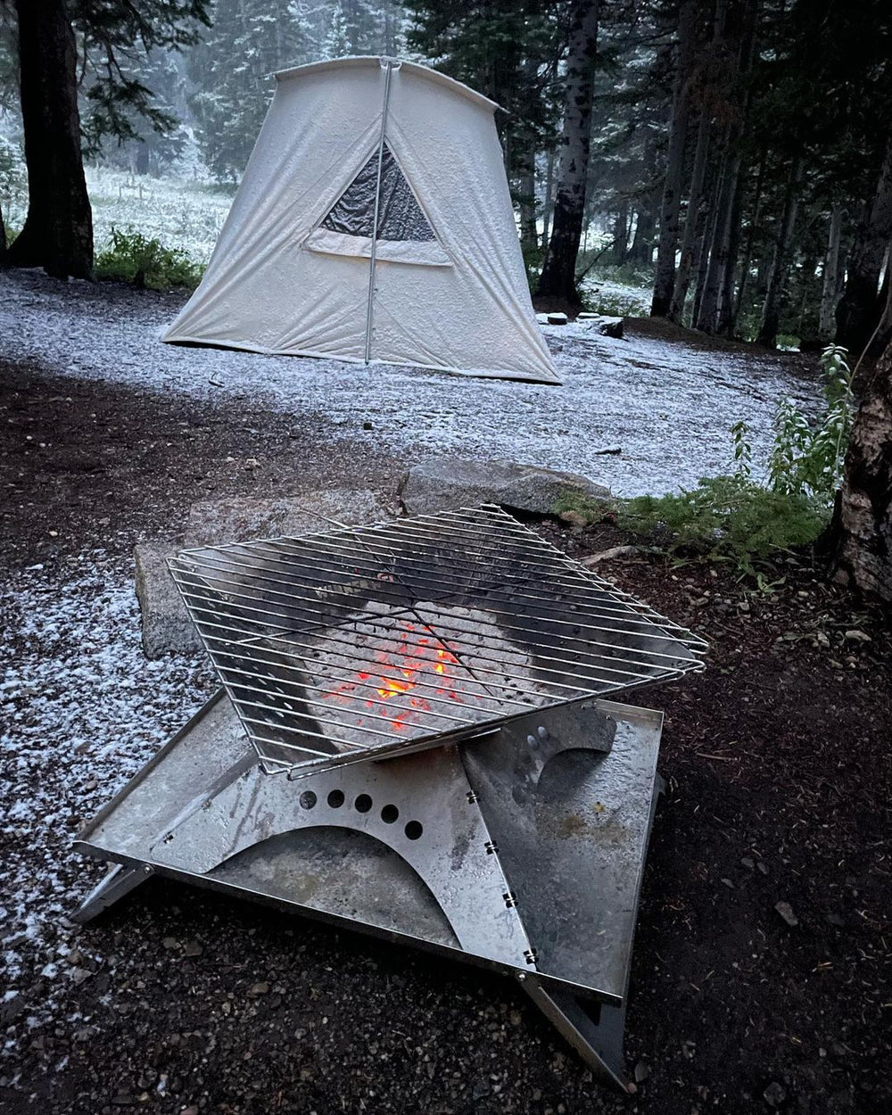 Foyer extérieur Winnerwell Flat avec grille de cuisson et braises dans la neige devant une tente en toile