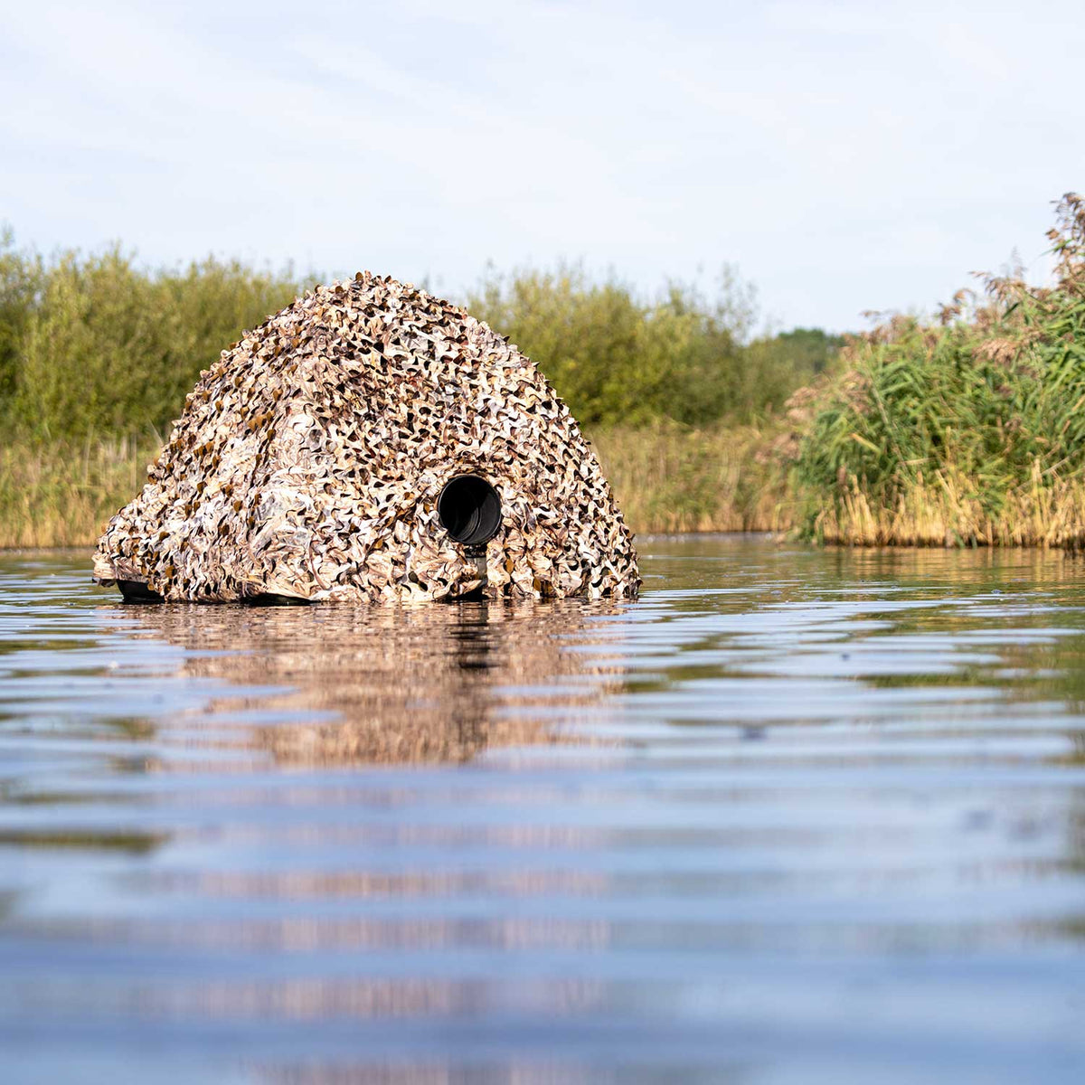 Filet de camouflage Buteo 4 sur un affût dans l'eau, avec une ouverture pour objectif et des roseaux sur la rive