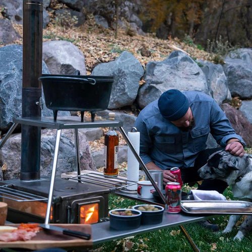 Winnerwell Fire Guard Stove Table près d'un poêle allumé avec casserole au-dessus et nourriture sur le rebord