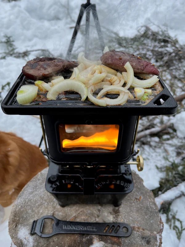 Winnerwell Iron Camping Cooker Stove dans la neige, viande et oignon sur la plaque et feu derrière la fenêtre