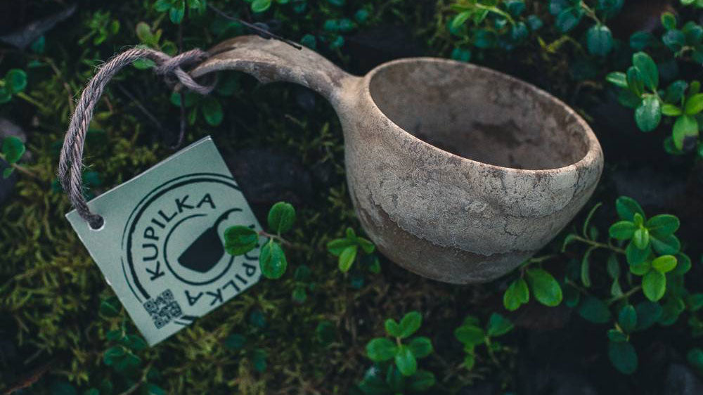 Kupilka, lichtgrijze kuksa met een touwtje en een label tussen groen mos