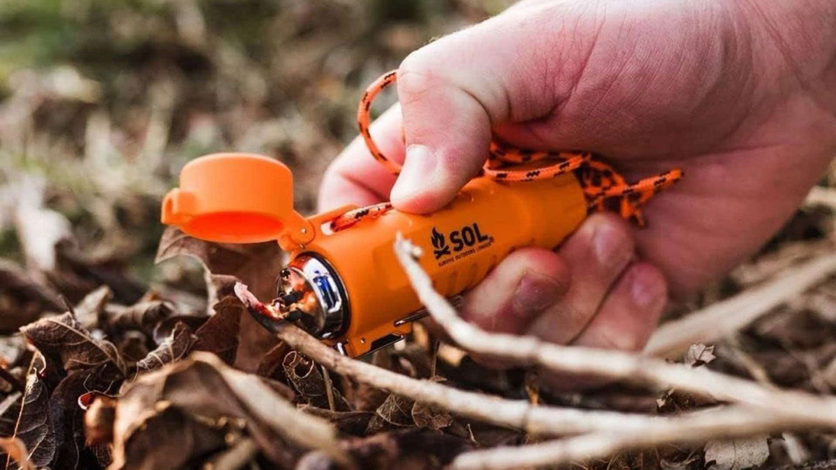 Aanstekers en vuurmakers, hand met een oranje vuurmaker aan een koord boven droge bladeren