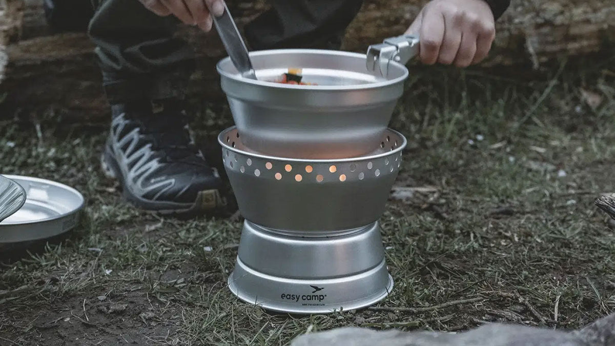 Pannensets, hand roert in een pan op een kampeerbrander met windscherm buiten op de grond