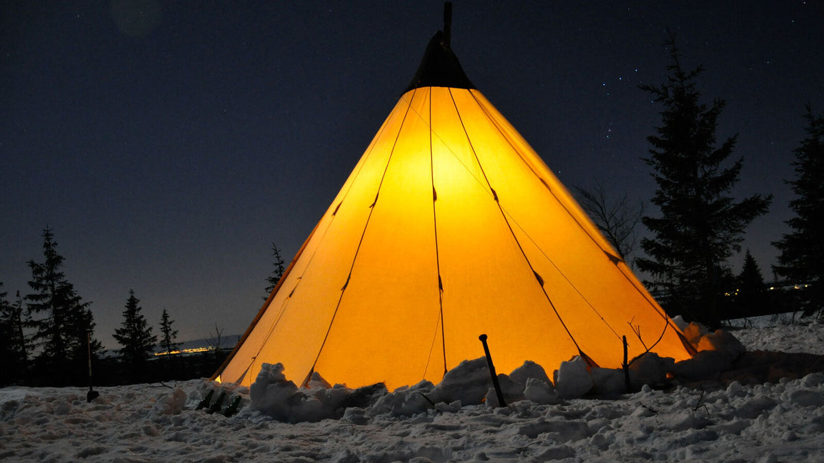 Tipi tent die van binnen oplicht in de sneeuw onder een sterrenhemel tussen dennen