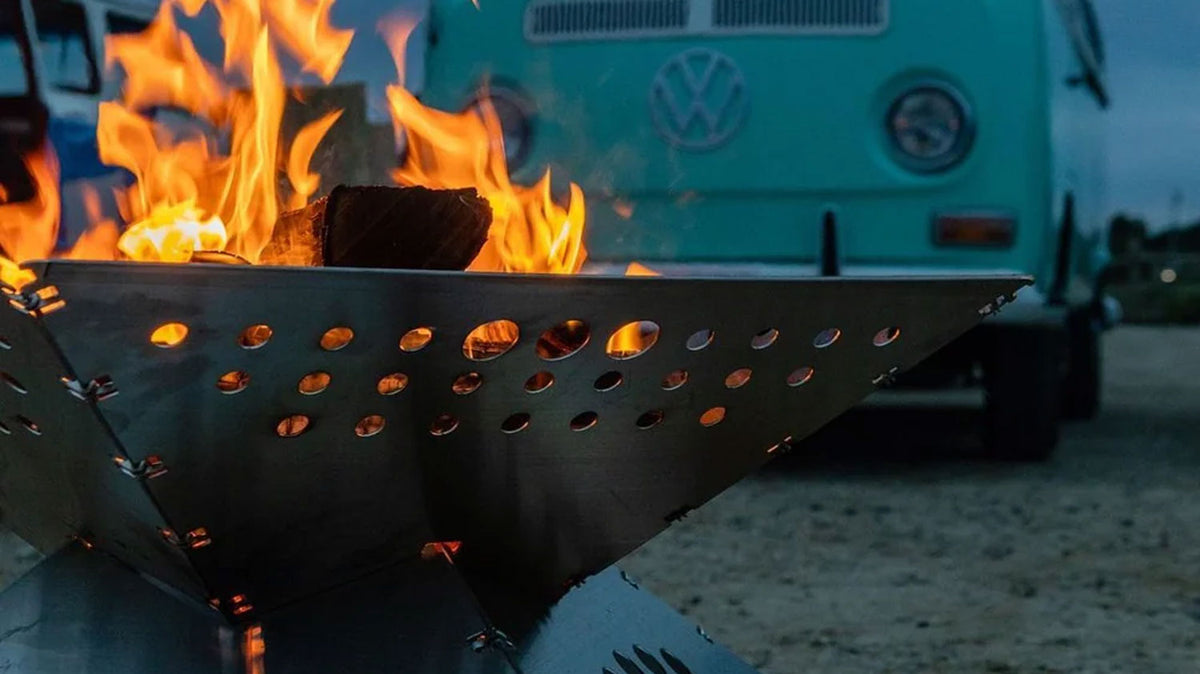 Vuurkorven met brandend hout in een metalen vuurkorf met gaatjes, met een mintgroen Volkswagen busje erachter