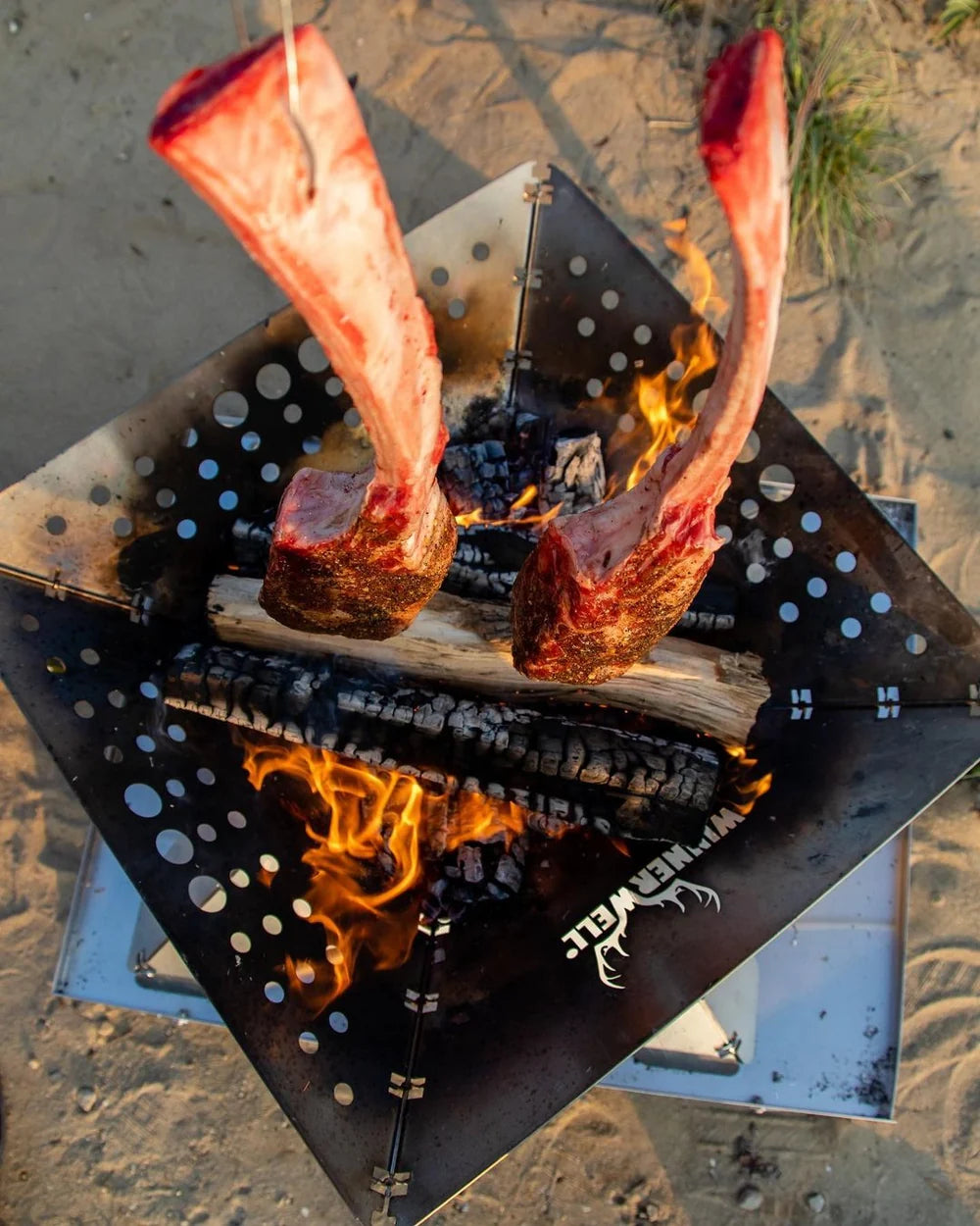 Foyer extérieur Winnerwell Flat vu d'en haut avec deux morceaux de viande sur l'os au-dessus du feu sur le sable
