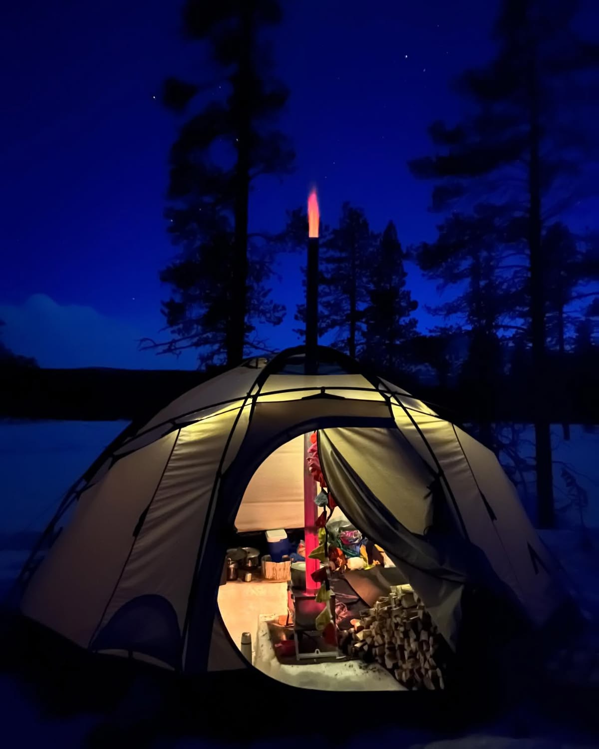 Tente dans la neige la nuit avec le Winnerwell Nomad View à l'intérieur et une flamme sortant de la cheminée