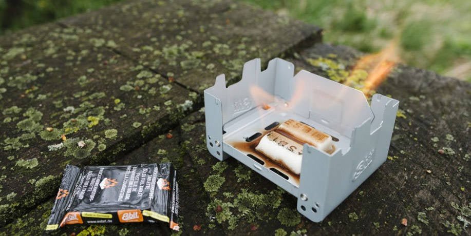 Esbit Survival Stove utilisé sur un muret de pierre moussu, avec flammes et un paquet de tablettes à côté