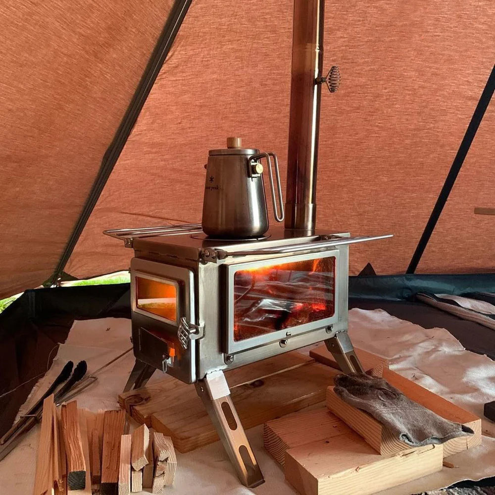 De Winnerwell Nomad View 1G houtkachel met dubbele kijkvensters, waarin een brandend vuur zichtbaar is. De kachel staat op een houten vloer in een tent en heeft een roestvrijstalen behuizing met een glazen zij- en voorpaneel. Bovenop de kachel staat een metalen koffiepot en er is een schoorsteenpijp aangesloten voor rookafvoer.