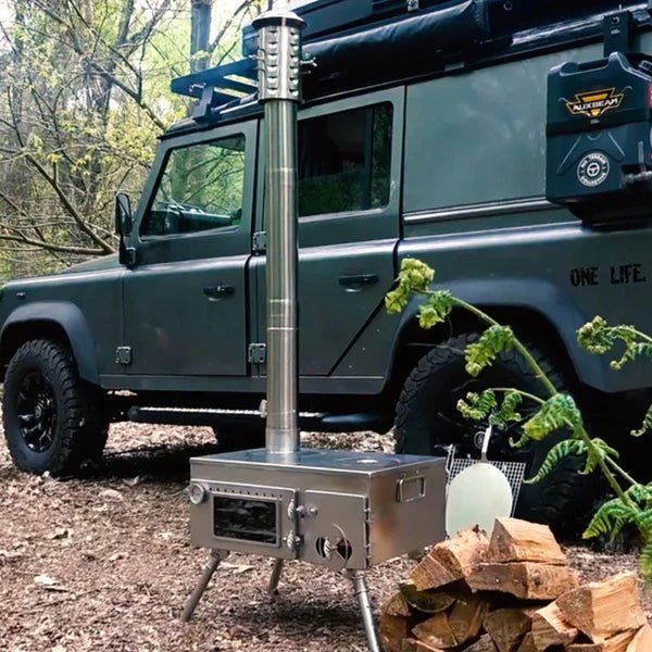 Winnerwell Woodlander Pizza Oven dehors en forêt devant un véhicule tout-terrain, avec du bois à côté